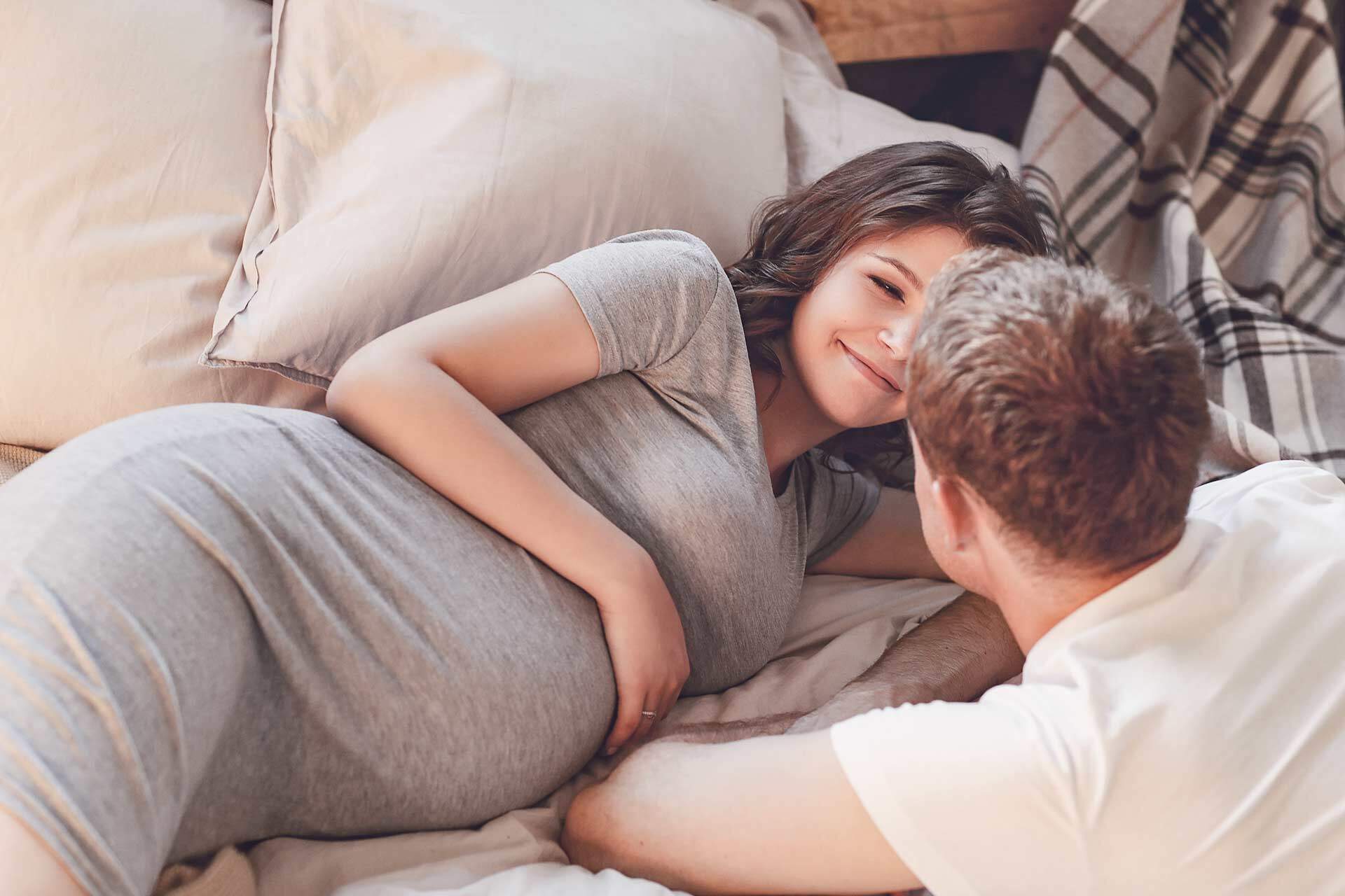 Bild zeigt eine liegende schwangere Frau auf dem Sofa. Ihr Partner beugt sich zu ihr. Sie lächelt.