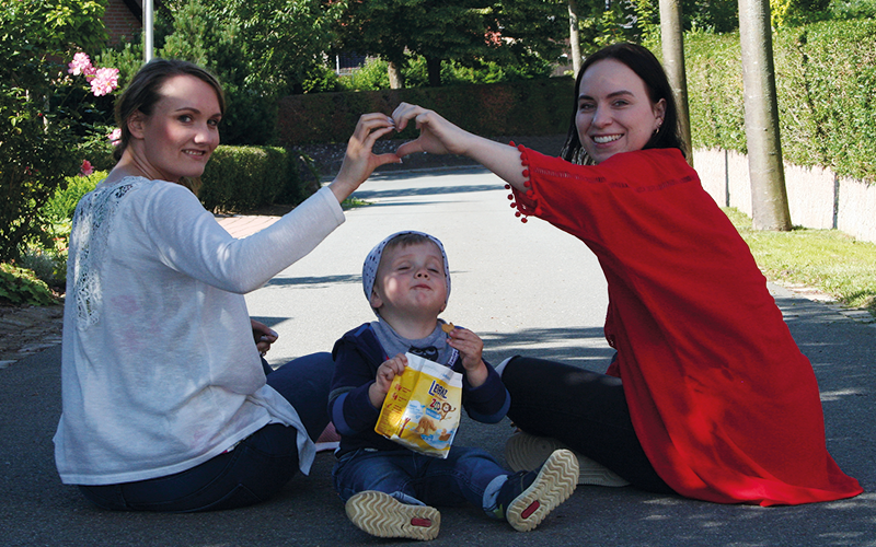 Lisa und Anika sitzen im schönen Sonnenlicht auf der Straße und formen über dem Kopf von Jonah aus einer Hand ein Herz.