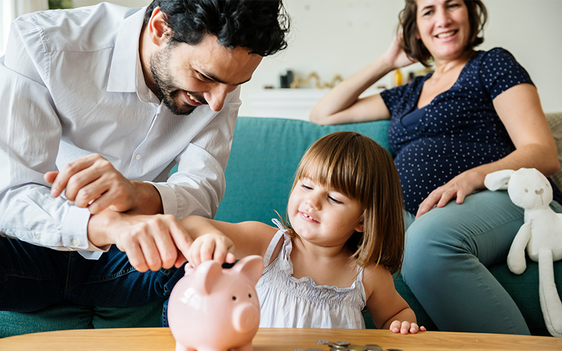 Vater und Tochter werfen Geld in ein Sparschwein. Die schwangere Mutter sitzt im Hintergrund auf dem Sofa und lächelt.